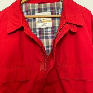Peut inclure: Une veste rouge avec une fermeture &eacute;clair et deux poches &agrave; rabat. La veste est doubl&eacute;e de plaid et porte une &eacute;tiquette "London Fog" avec le texte "LONDON, ENGLAND, NEW YORK, TRADEMARK OF LONDON FOG CORPORATION, MADE IN TURKEY".