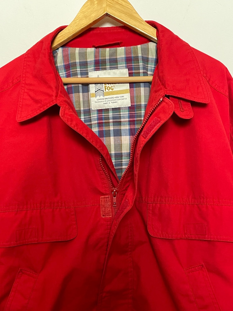 Peut inclure: Une veste rouge avec une fermeture &eacute;clair et deux poches &agrave; rabat. La veste est doubl&eacute;e de plaid et porte une &eacute;tiquette "London Fog" avec le texte "LONDON, ENGLAND, NEW YORK, TRADEMARK OF LONDON FOG CORPORATION, MADE IN TURKEY".