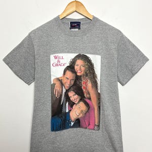 May include: A gray t-shirt featuring a graphic of the cast of the television show "Will & Grace." The graphic shows four people, with the show's title in white text. The shirt is hanging on a wooden hanger.