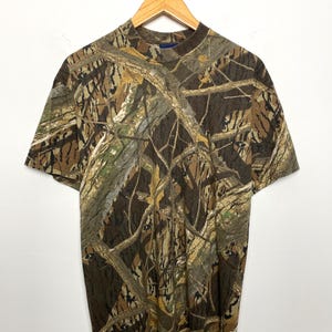 May include: A short-sleeved t-shirt with a camouflage pattern in shades of brown, green, and tan. The shirt is displayed on a wooden hanger against a white backdrop. The camouflage design is reminiscent of a woodland setting.
