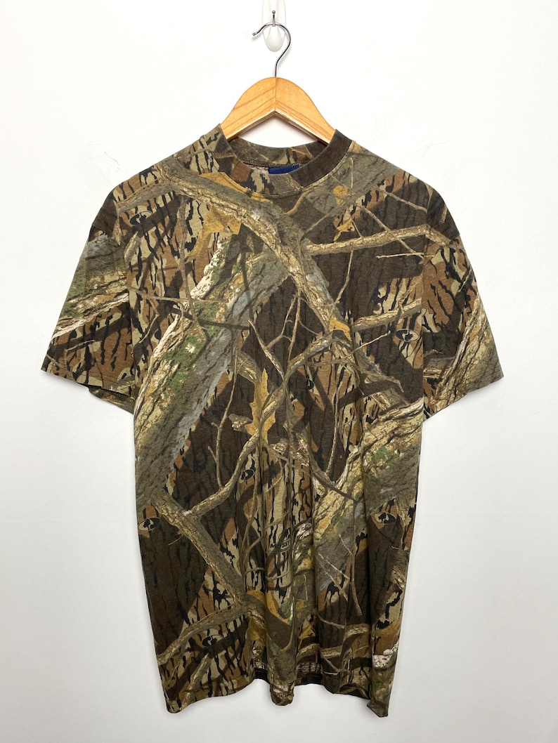 May include: A short-sleeved t-shirt with a camouflage pattern in shades of brown, green, and tan. The shirt is displayed on a wooden hanger against a white backdrop. The camouflage design is reminiscent of a woodland setting.
