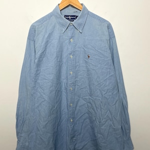May include: Light blue button-down shirt with long sleeves, hanging on a wooden hanger. The shirt features a button-down collar, a small embroidered logo on the chest, and the brand name on the collar.