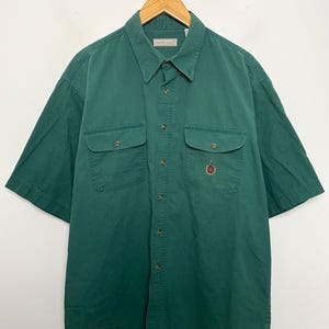 May include: A short-sleeved button-down shirt in a solid dark green color. The shirt features a pointed collar, a front button placket with brown buttons, and two chest pockets with buttoned flaps. An embroidered crest logo sits on the left pocket.