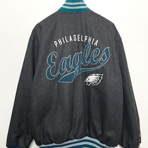 May include: A gray bomber jacket with a teal and white striped collar and cuffs. The back of the jacket features the words "Philadelphia Eagles" in a white and teal script font with an embroidered eagle logo.