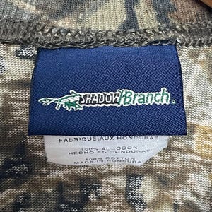 May include: A close-up of a camouflage-patterned shirt. A navy blue tag displays the text "SHADOW BRANCH" in white and green. Below the tag, a white label reads "FABRIQUE AUX HONDURAS" and "100% COTTON MADE IN HONDURAS".