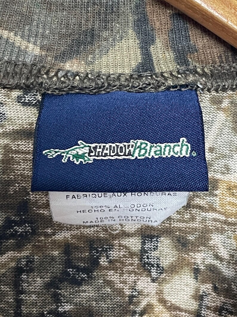 May include: A close-up of a camouflage-patterned shirt. A navy blue tag displays the text "SHADOW BRANCH" in white and green. Below the tag, a white label reads "FABRIQUE AUX HONDURAS" and "100% COTTON MADE IN HONDURAS".