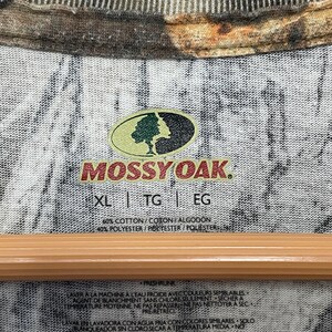 May include: Close-up of a camouflage-patterned shirt with the "Mossy Oak" logo. The shirt's tag indicates sizes XL, TG, and EG, with fabric composition details. The shirt has a crew neck and a wooden hanger.