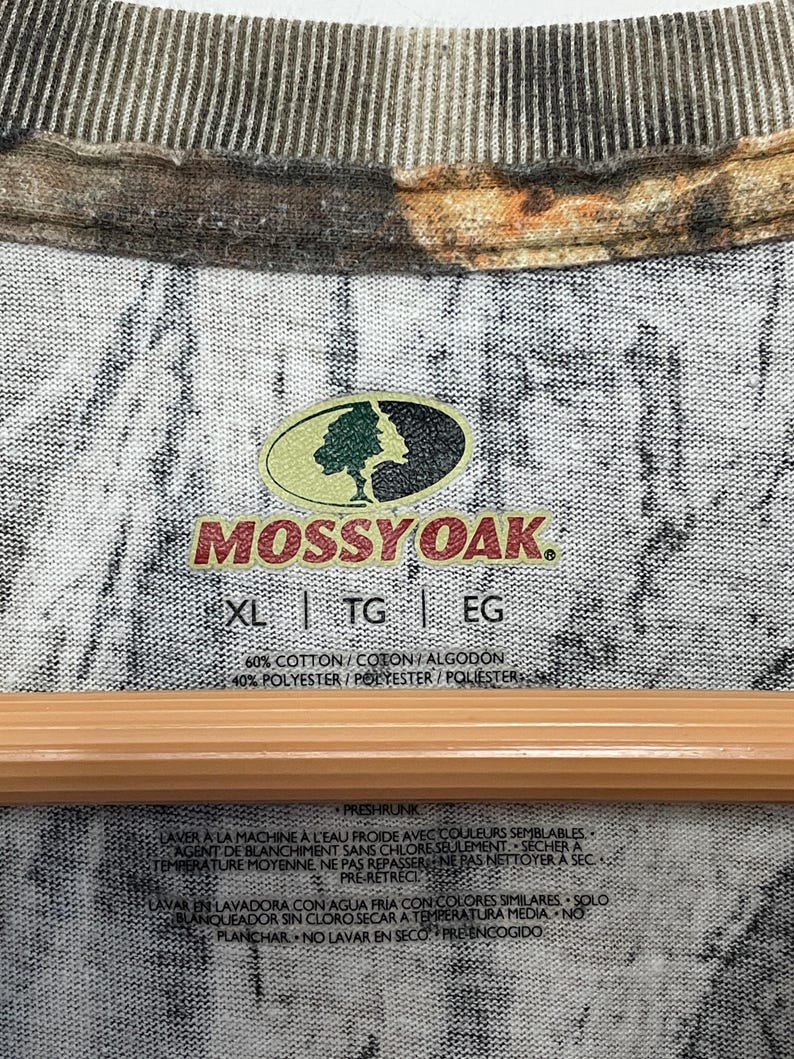May include: Close-up of a camouflage-patterned shirt with the "Mossy Oak" logo. The shirt's tag indicates sizes XL, TG, and EG, with fabric composition details. The shirt has a crew neck and a wooden hanger.
