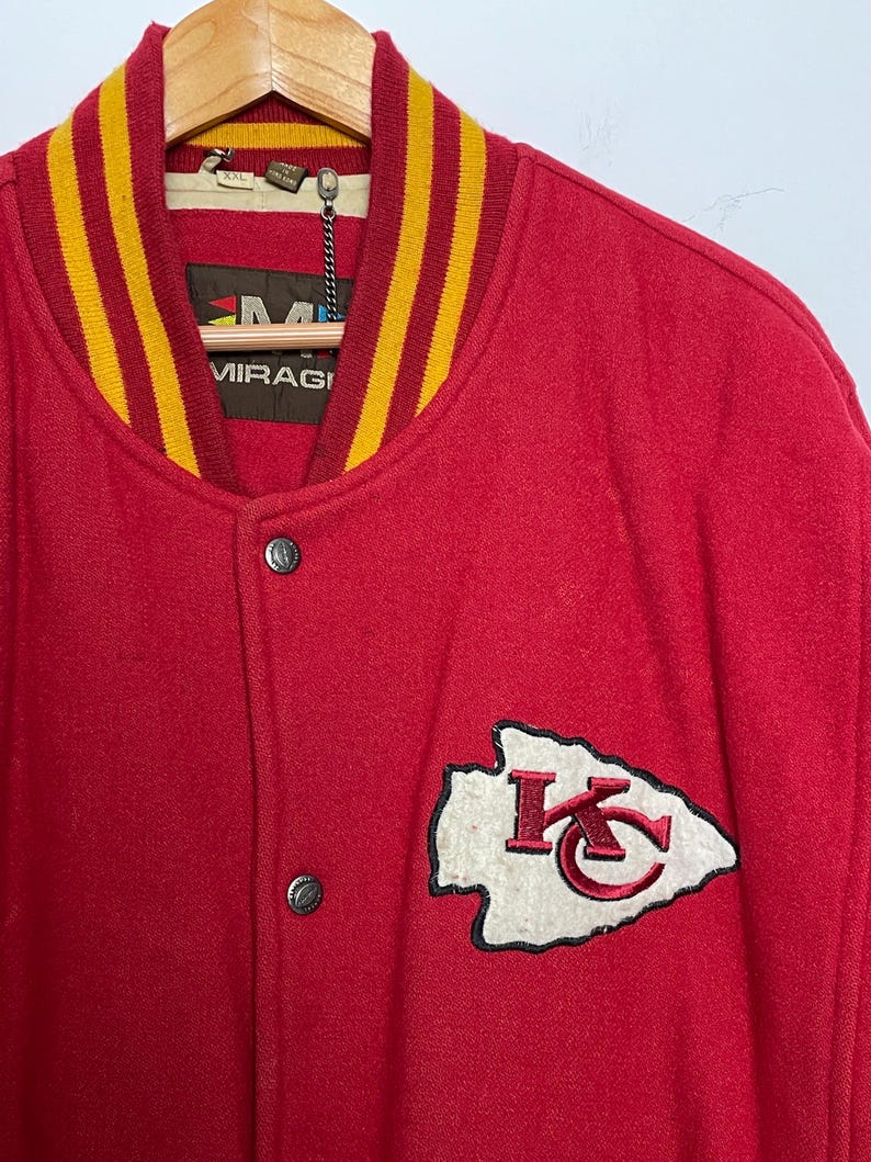 May include: Red wool varsity jacket with a yellow and gold striped collar and cuffs. Features a white and red embroidered team logo patch on the chest. The jacket has silver snap closures and a tag that reads "MIRAGI".