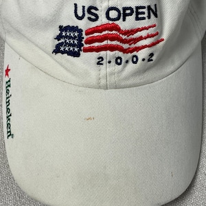 May include: A white baseball cap featuring embroidered text that reads US OPEN 2.0.0.2 with a stylized American flag graphic. The brim of the cap displays the green text Heineken accompanied by a small red star symbol on the left side.
