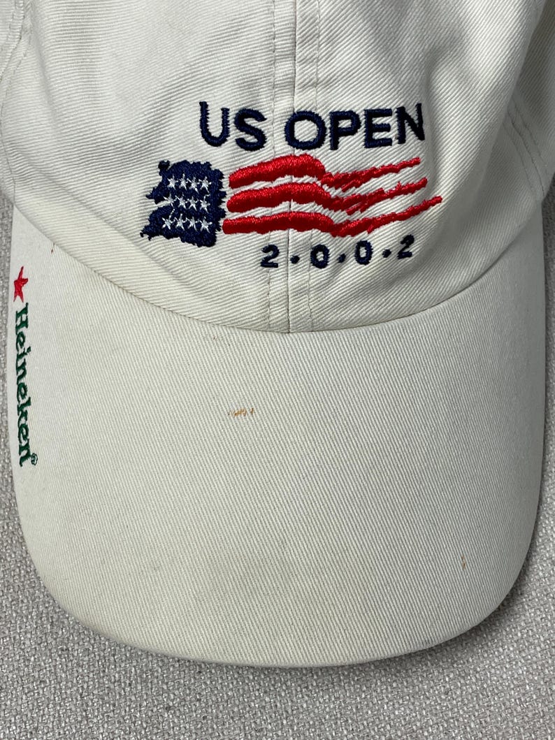 May include: A white baseball cap featuring embroidered text that reads US OPEN 2.0.0.2 with a stylized American flag graphic. The brim of the cap displays the green text Heineken accompanied by a small red star symbol on the left side.