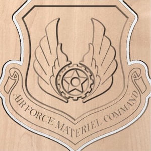 Può includere: Targa in legno con l'emblema dell'Air Force Materiel Command. L'emblema ha la forma di uno scudo con ali, un ingranaggio e una stella al centro. Le parole "AIR FORCE MATERIEL COMMAND" sono incise su uno stendardo attorno allo scudo.