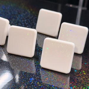 Puede incluir: Primer plano de varios objetos blancos de forma cuadrada con bordes redondeados. Cada objeto tiene un sutil efecto de purpurina iridiscente. Los objetos están dispuestos sobre una superficie oscura y moteada.