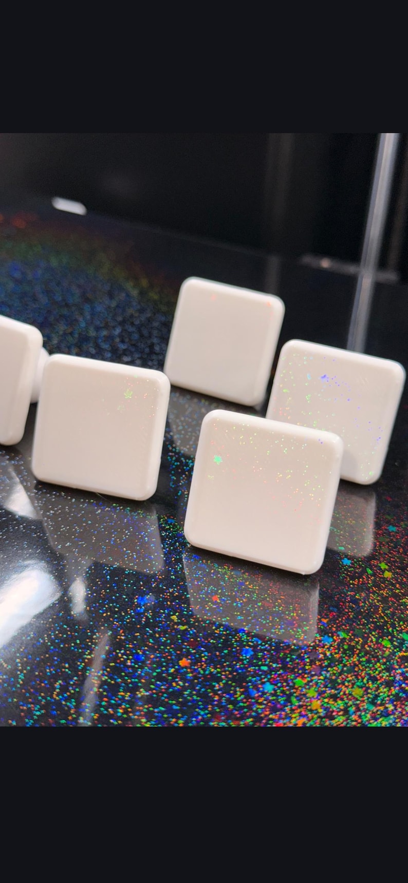 Pode incluir: Close-up de v&aacute;rios objetos brancos em forma de quadrado com bordas arredondadas. Cada objeto tem um efeito sutil de glitter iridescente. Os objetos est&atilde;o dispostos sobre uma superf&iacute;cie escura e salpicada.