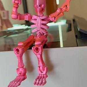 Puede incluir: Una figura de esqueleto articulada rosa y naranja. El esqueleto impreso en 3D está posado con los brazos levantados. El degradado de color pasa del rosa al naranja en los brazos y las piernas. La figura está sentada sobre una superficie blanca.