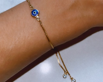 Può includere: Bracciale dorato con un ciondolo a forma di occhio diabolico con smalto blu e bianco. La catena regolabile include due piccoli ciondoli rotondi e trasparenti. Un gioiello delicato.