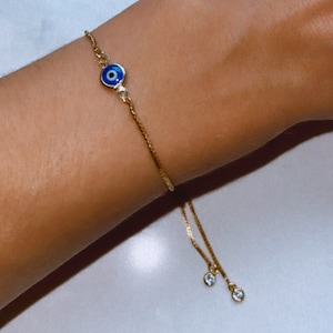 May include: Gold-toned bracelet featuring an evil eye charm with blue and white enamel. The adjustable chain includes two small, clear, round charms. A delicate piece of jewelry.