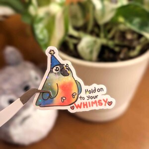 May include: A colorful sticker of a bird wearing a blue party hat, with the text "Hold on to your WHIMSY". The bird has a red belly, blue wings, and a yellow star on its head. The sticker is held by tweezers.