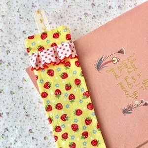 May include: A yellow fabric bookmark with a red and white polka dot ruffle and a strawberry print. The bookmark has a loop at the top for hanging.