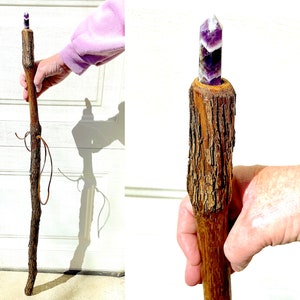 May include: A walking stick with a purple amethyst crystal at the top. The stick is made of natural wood with a brown bark-like texture. The stick has a leather strap wrapped around it.