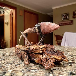 May include: A handcrafted decorative sculpture featuring a pink arrowhead mounted on a weathered wooden base. The arrowhead is supported by metal nails and a section of antler. The base has a detailed, textured appearance.