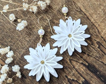 May include: A pair of white daisy-shaped earrings with gold-colored ear wires and a small pearl bead. The center of each flower has a gold-colored accent. The earrings are displayed on a wooden surface.
