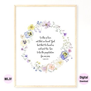 May include: A watercolor floral wreath with purple, yellow, and blue flowers surrounds a quote from the Bible, 1 John 4:10, in a white frame. The quote reads, "In this is love, not that we loved God, but that He loved us and sent His Son to be the propitiation for our sins." The text is in black and the background is white.