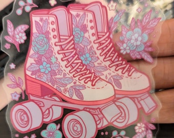 May include: Pink roller skates with floral designs and white wheels. The skates have pink laces and are decorated with pink, blue, and white flowers.