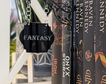 May include: Black metal sign with the word "FANTASY" hanging from a decorative chain. The sign is in front of a row of books with black covers. The sign is attached to a decorative metal piece that sits on top of the books.
