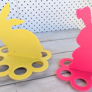 May include: Two Easter bunny egg holders, one yellow and one pink, with a flower-shaped base and six egg-shaped slots.