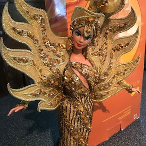 May include: A gold Barbie doll wearing a gold dress and a gold crown with a sun design. The doll is standing in front of a box with the word "Sun" on it.