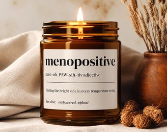 Puede incluir: Una vela encendida en un frasco de vidrio ámbar con una etiqueta blanca que presenta el texto menopositive, una pronunciación fonética y la definición: Finding the bright side in every temperature swing. See also: empowered, upbeat.