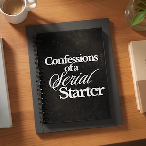 Op de afbeelding: Een zwart spiraalgebonden notitieboek met de titel "Confessions of a Serial Starter" in witte letters. Het notitieboek ligt op een grijze ondergrond. Een kop koffie en een boek zijn zichtbaar op de achtergrond.