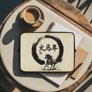 May include: A laptop sleeve featuring a black and white illustration of a running horse, with Chinese characters inside a black circle. The sleeve is on a white surface with a cup of coffee, a notepad, and a pen.