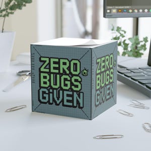 Op de afbeelding: Een grijze, kubusvormige notitieblok met een witte sticky note erop. De notitieblok heeft een rasterpatroon en de tekst "ZERO BUGS GIVEN" in groene en zwarte gepixelde letters.