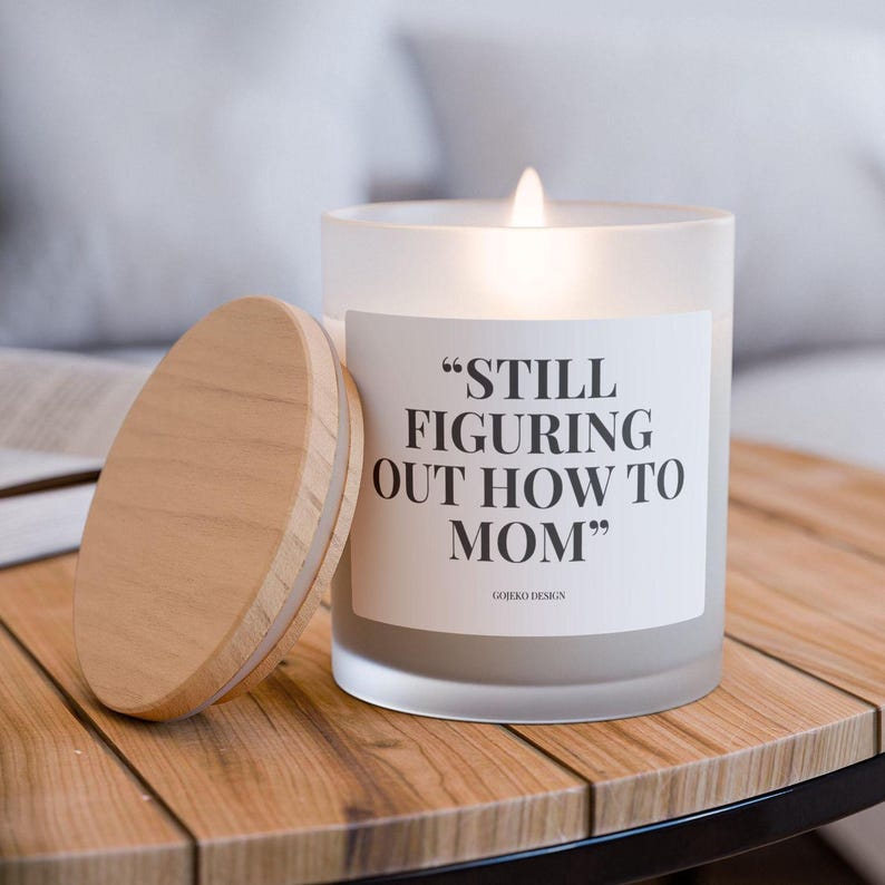 Aesthetic frosted glass candle with a wooden wick and a humorous label reading "Still Figuring Out How to Mom." Perfect for new moms, seasoned moms, and moms in need of self-care. Made from a coconut soy wax blend with a crackling wick for a cozy ambiance. A thoughtful, funny mom gift ideal for Mother’s Day, baby showers, or just because. Comes in a chic frosted jar with a raw maple lid. A stylish and practical addition to any mom’s space, blending humor with relaxation.