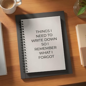 May include: A spiral-bound notebook with the text "THINGS I NEED TO WRITE DOWN SO I REMEMBER WHAT I FORGOT" on the open page. The notebook is on top of a larger, dark gray notebook. A cup of coffee and a book are in the background.