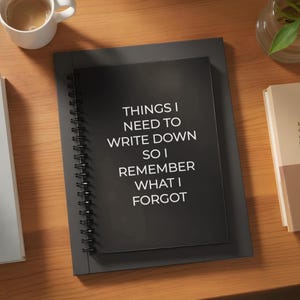 May include: A black spiral-bound notebook with the text "THINGS I NEED TO WRITE DOWN SO I REMEMBER WHAT I FORGOT" in white. The notebook is on top of a larger, black, rectangular notebook. A cup of coffee and a book are in the background.