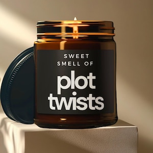 May include: A brown glass candle jar with a black label that reads "Sweet Smell of Plot Twists". The candle is lit and the jar has a gold lid.