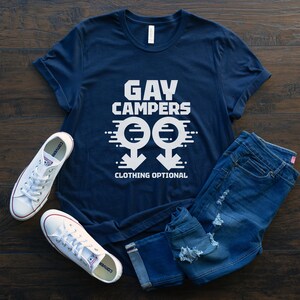 May include: Navy blue t-shirt with the text "GAY CAMPERS CLOTHING OPTIONAL" in white. The shirt is paired with blue jeans and white sneakers with red accents. The design includes two gender symbols.