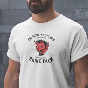 May include: White t-shirt featuring a red devil graphic with the text "THE DEVIL WHISPERED I'M COMING FOR YOU, I WHISPERED BACK... BRING DICK". The devil graphic has a red face, horns, and a mustache.