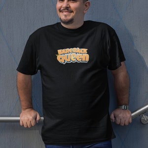 May include: Black t-shirt with the text "Bareback Queen" in orange and yellow. The shirt is worn by a person in blue jeans. The background is a gray wall with blue diamond-shaped lines.