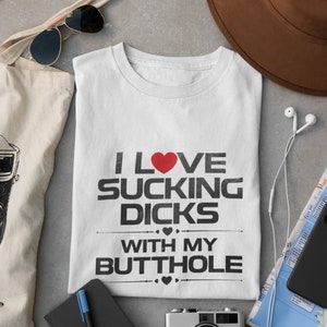 May include: White t-shirt with the text "I LOVE SUCKING DICKS WITH MY BUTTHOLE" in black font and a red heart. The shirt is surrounded by a brown hat, sunglasses, a tote bag, a notebook, a camera, and a map.