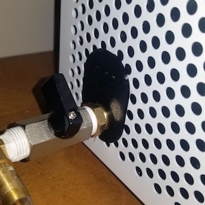 Puede incluir: Primer plano de una válvula negra con una manija plateada unida a un panel de metal blanco con una cubierta circular negra. El panel tiene una cuadrícula de pequeños agujeros.