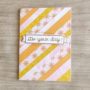 Puede incluir: Tarjeta de felicitación rectangular con un diseño de rayas diagonales en amarillo, rosa y blanco. La tarjeta presenta el texto "it's your day!" en una pancarta. Colocada sobre una superficie de madera.