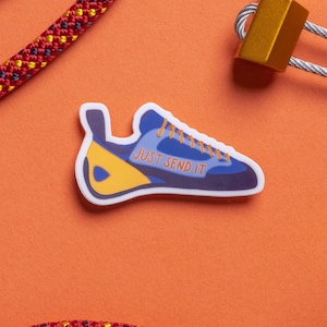 Puede incluir: Una pegatina de zapato de escalada azul y amarillo con el texto "JUST SEND IT" sobre un fondo naranja con cuerda de escalada roja y amarilla y equipo de escalada.