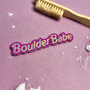 Peut inclure: Un patch thermocollant violet et pailleté avec le texte "Boulder Babe" en paillettes arc-en-ciel.