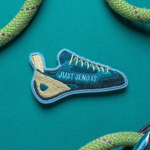 May include: A blue and tan embroidered patch in the shape of a climbing shoe, with the text "JUST SEND IT" in white. The patch is on a teal background, with green and tan climbing rope in the background.