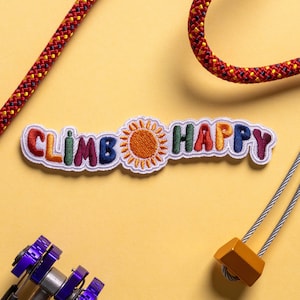 Pode incluir: Um remendo bordado colorido com o texto "Climb Happy" e um sol amarelo no centro. O remendo está sobre um fundo amarelo com uma corda vermelha e amarela e um mosquetão de escalada prateado.