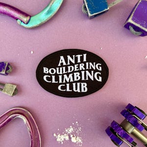 Può includere: Una toppa ovale nera con la scritta bianca "ANTI BOULDERING CLIMBING CLUB". L'attrezzatura da arrampicata in vari colori, tra cui viola, blu e turchese, circonda la toppa su uno sfondo lilla chiaro.